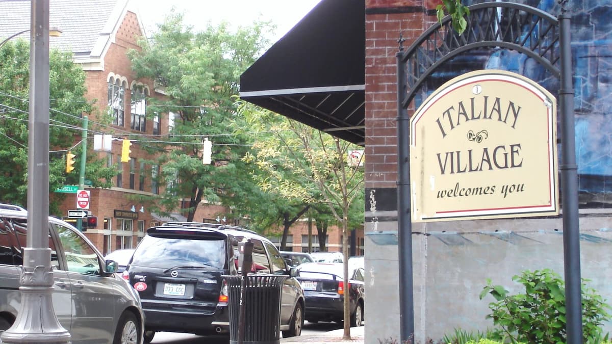 Archival photograph of an Italian Village neighborhood welcome sign beside trees and a Columbus street.