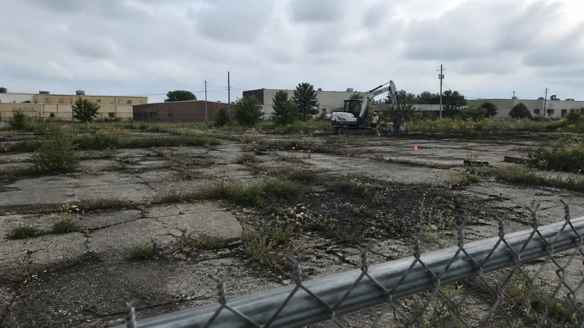 AI-generated illustration of cracked pavement, weeds and an excavator at an anonymous industrial parcel.