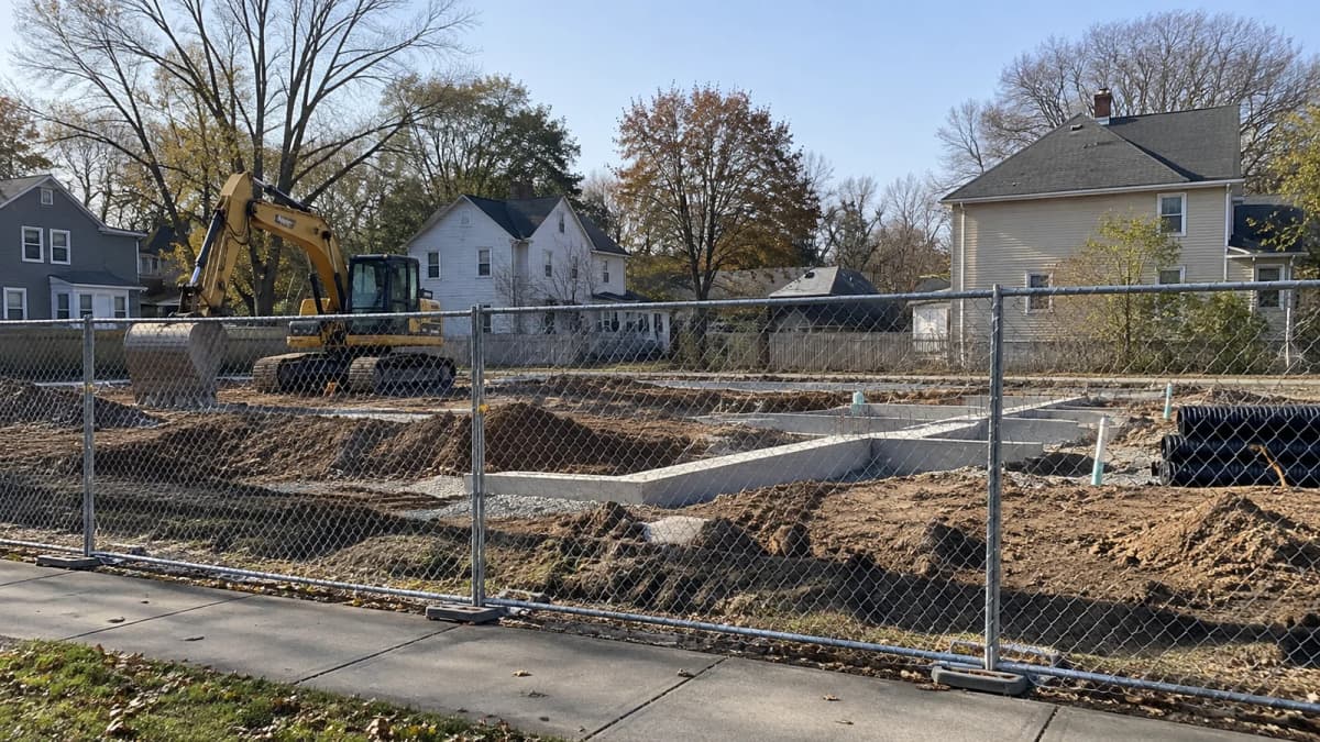 AI-generated illustration of foundation work and an excavator behind fencing beside neighborhood houses.