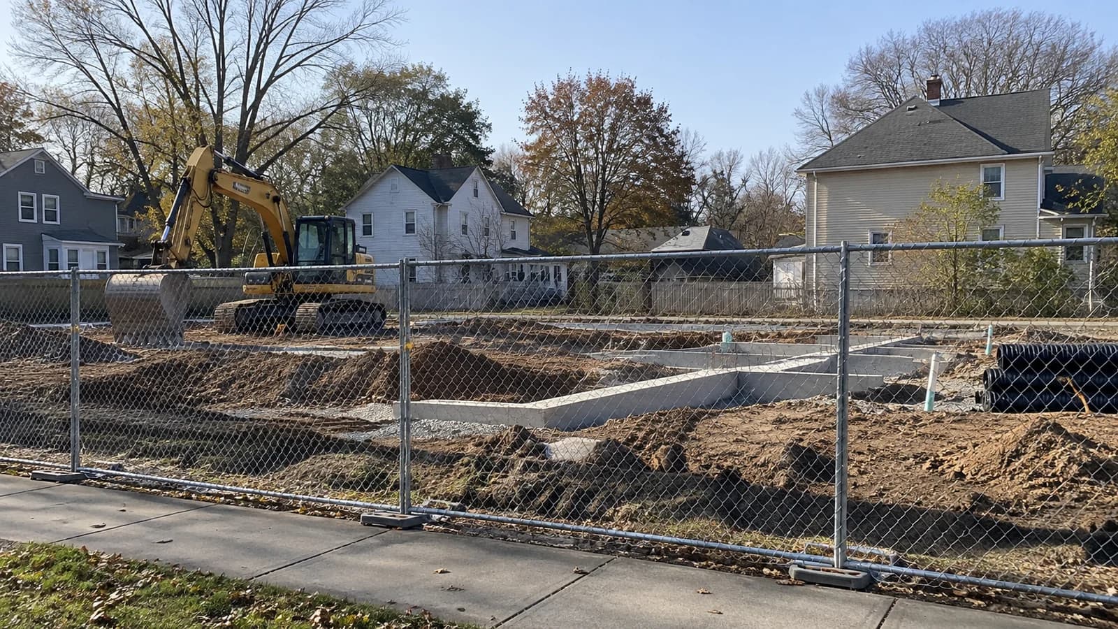 AI-generated illustration of foundation work and an excavator behind fencing beside neighborhood houses.