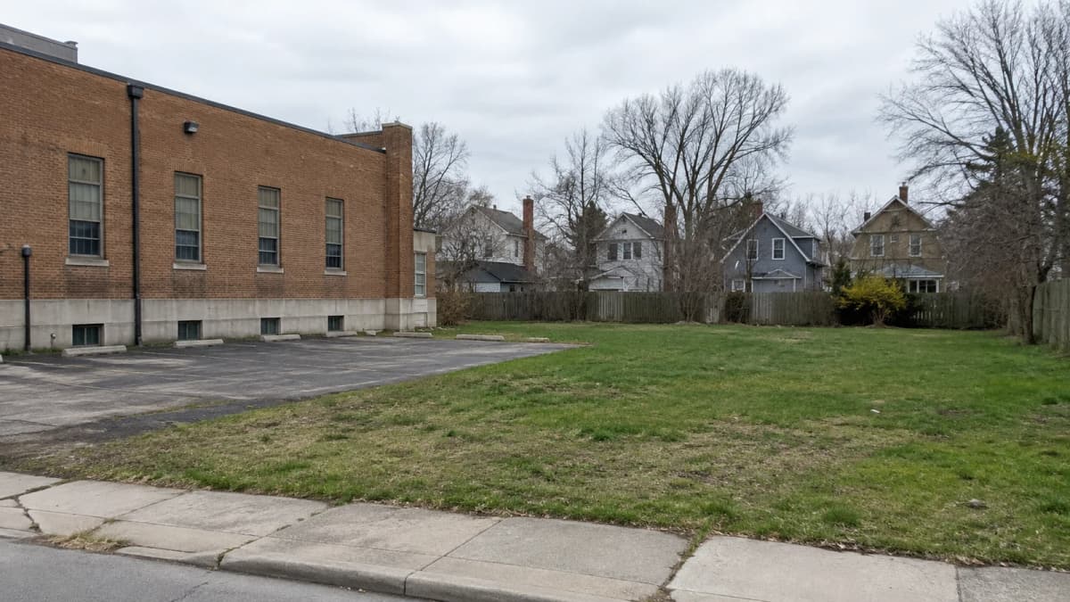 AI-generated illustration of a grassy vacant lot beside a brick building and older houses.
